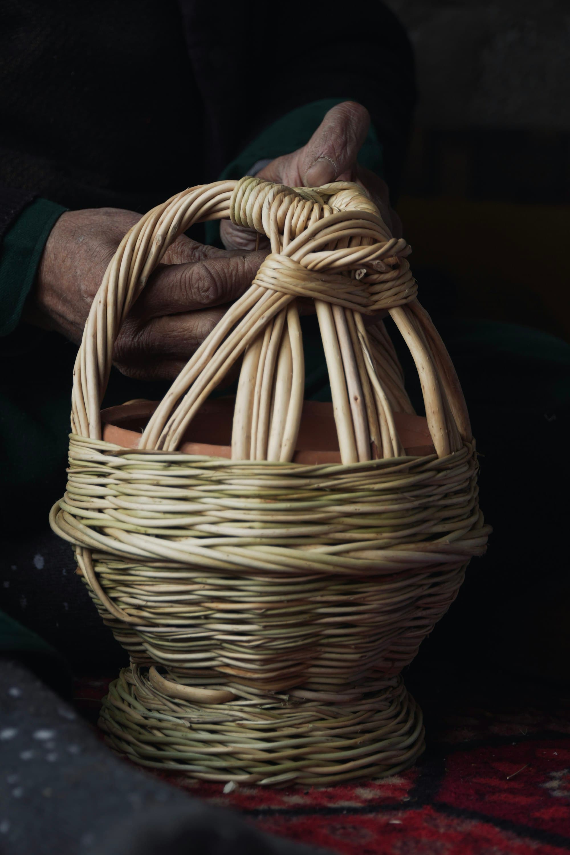 A maker’s hands lifting a half-finished willow basket from a dark workbench.