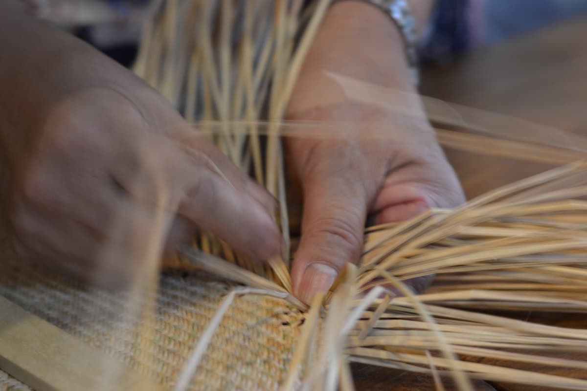 Hands weaving damp straw rods into the wall of a basket on a workbench.