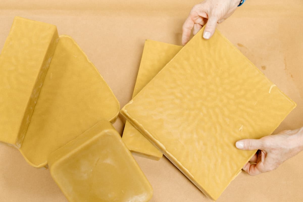 Hands lifting flat cakes of unbleached beeswax from a paper-lined table.