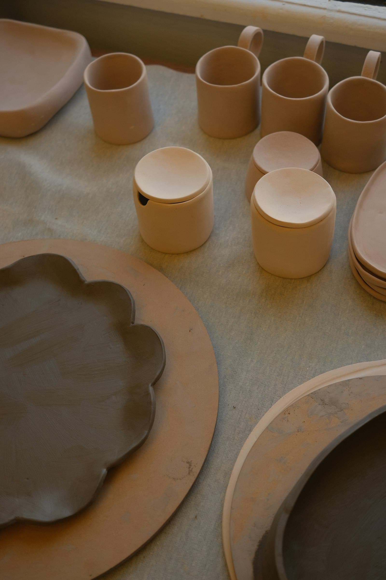 Unglazed clay candle holders and dishes drying on a canvas-covered bench.